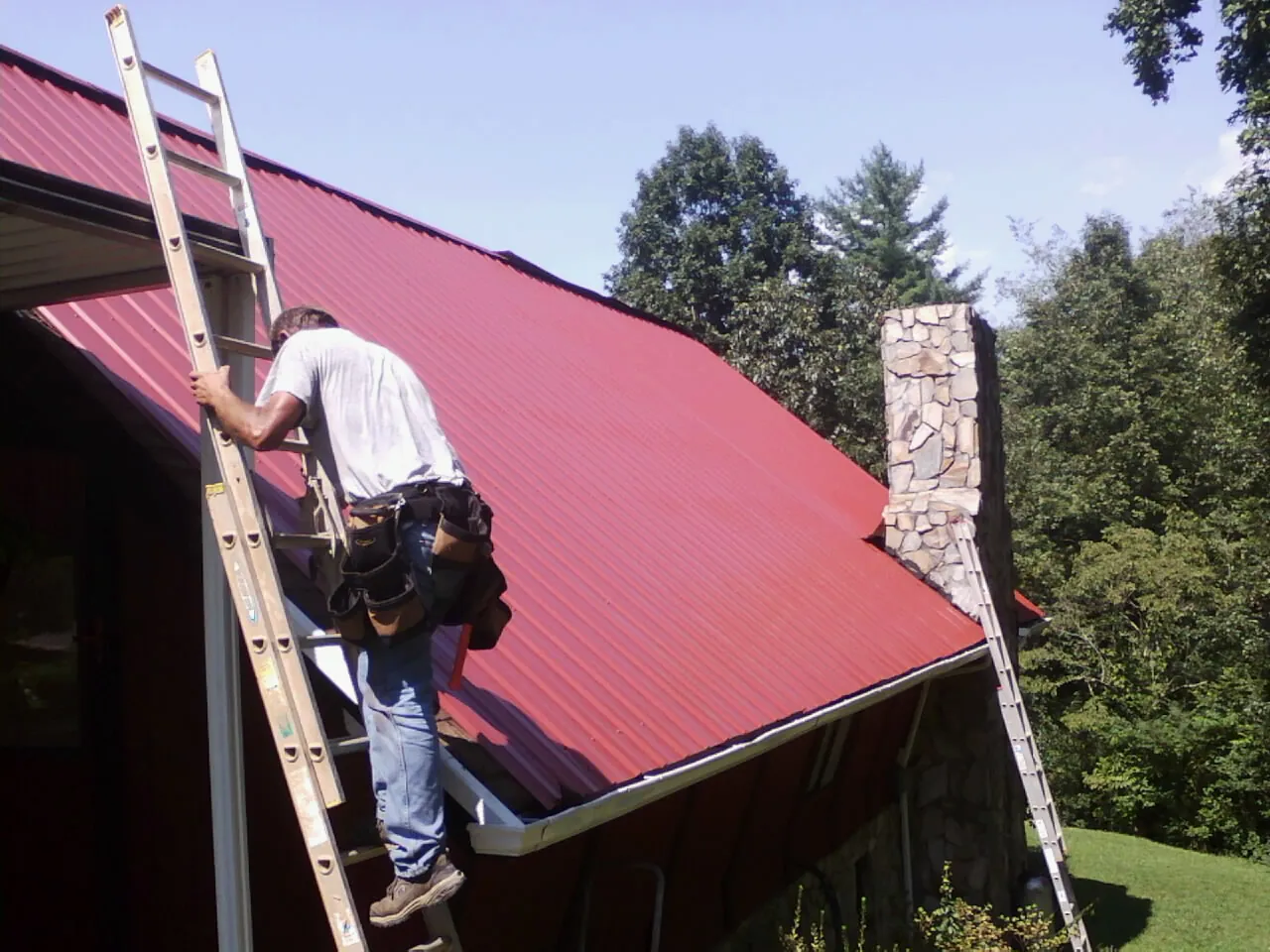 Roofer on a ladder beside a red metal roof and stone chimney — Soffit & Fascia Repair by Roofing Brothers in Piqua